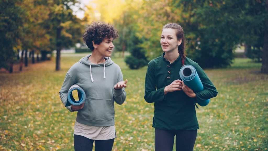Pretty girls are walking in park holding yoga mats and talking discussing sports equipment, smiling and laughing. Recreation, active lifestyle and autumn nature concept.